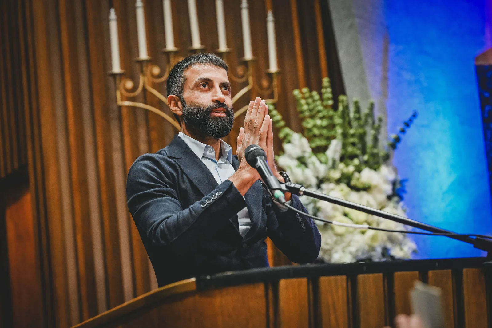 A bearded man in a suit speaks at a podium microphone with hands pressed together, a menorah behind him