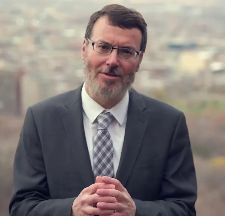 A bearded man in glasses and a grey suit smiles with hands clasped, outdoors