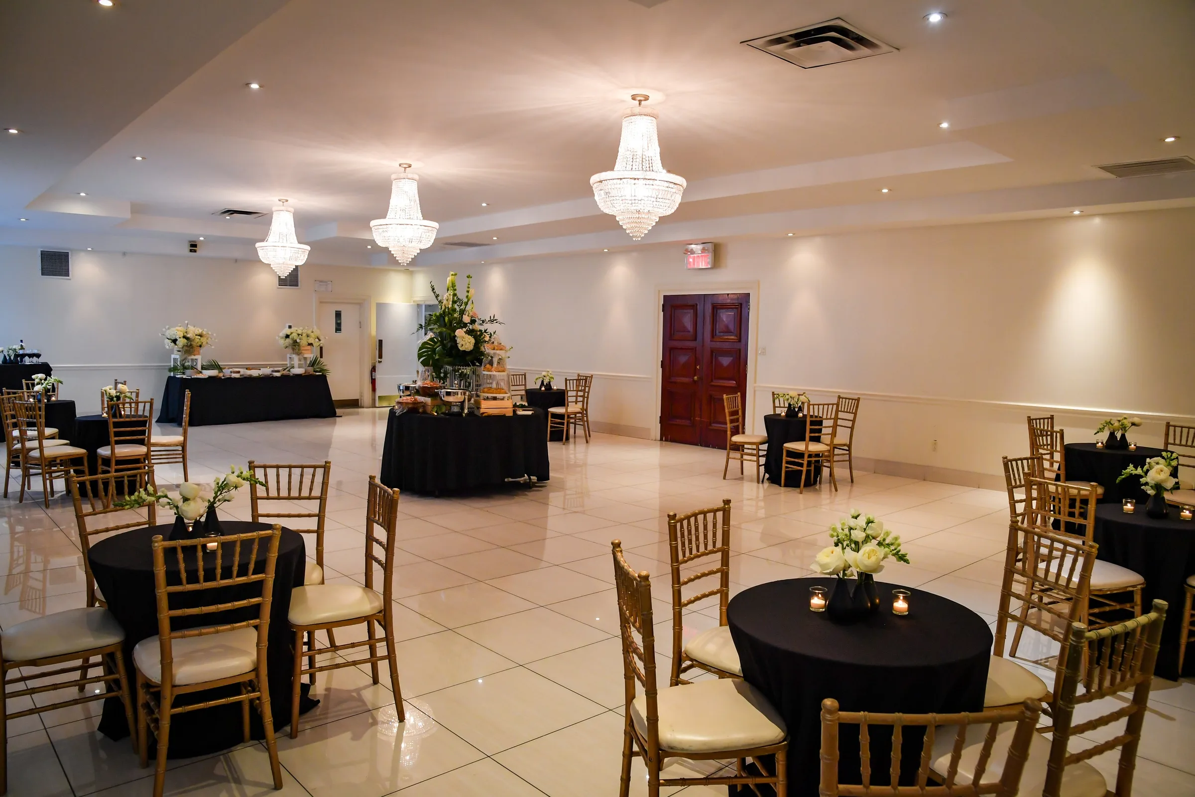 A white reception hall with crystal chandeliers, a tiled floor, wooden doors and round tables with gold chairs and white flowers