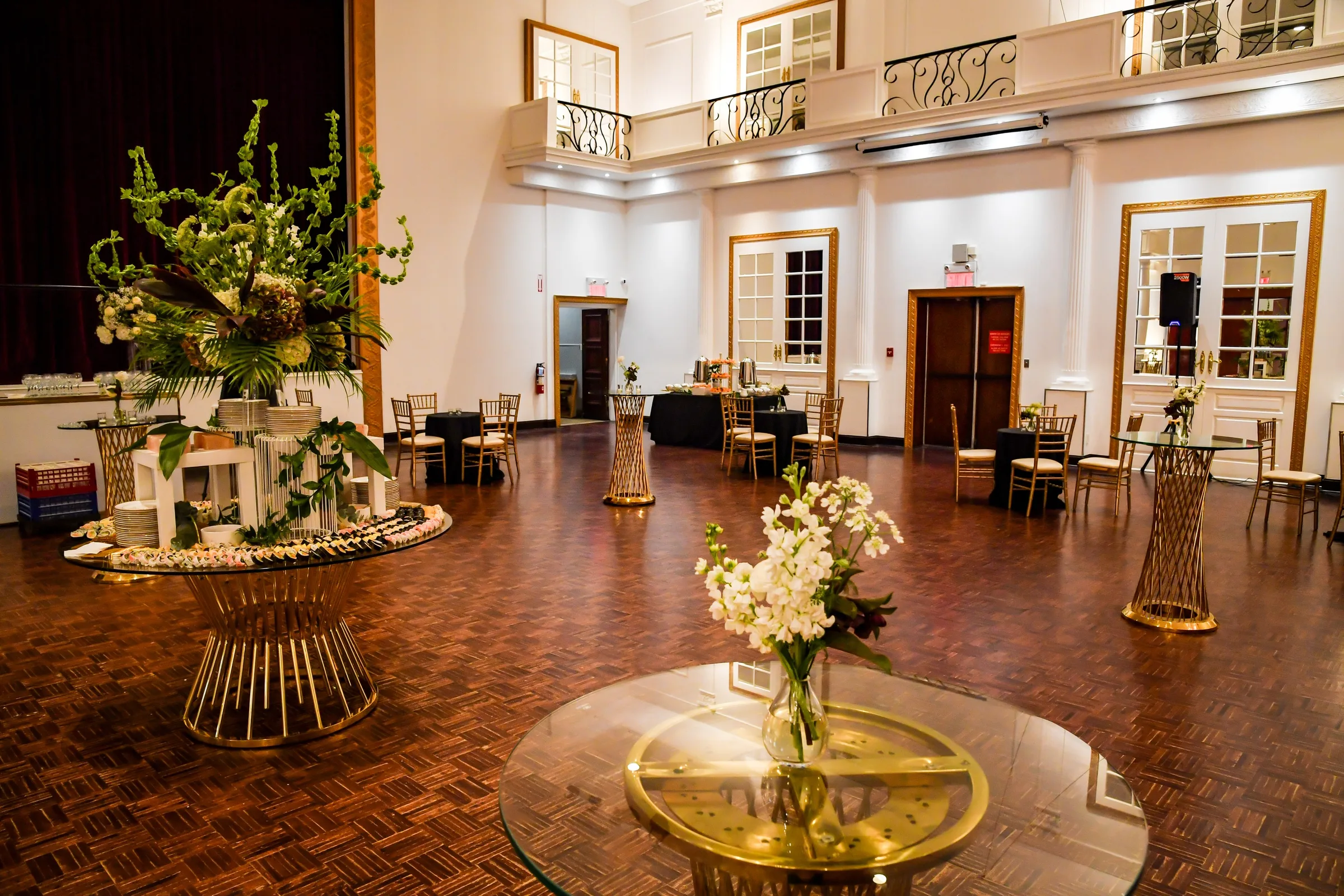 Singerman Hall set for a reception: Juliet balconies, French doors with gold frames, a parquet floor and tall flower arrangements