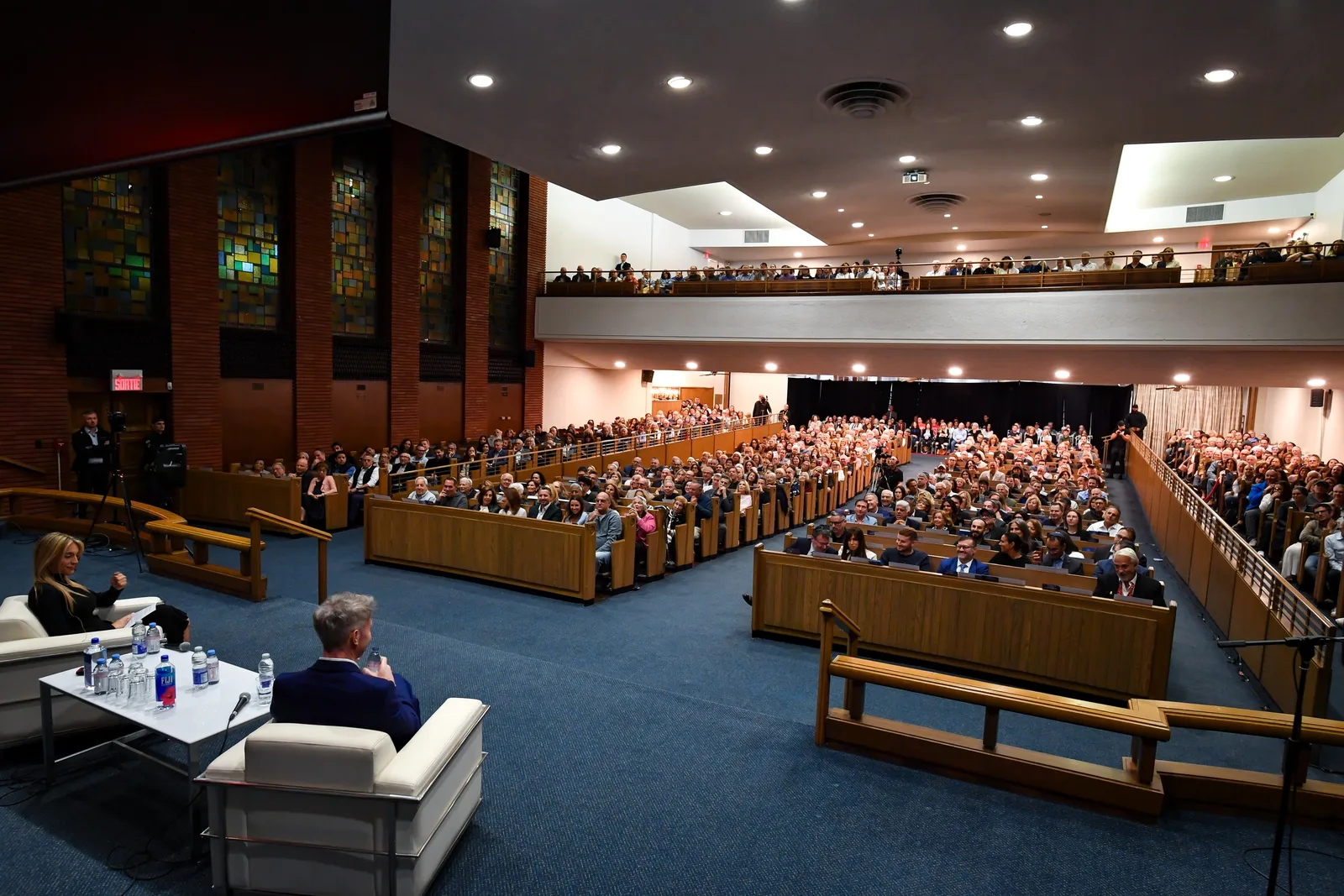 Two people in conversation in white armchairs at the front of the sanctuary, the full pews and balcony listening